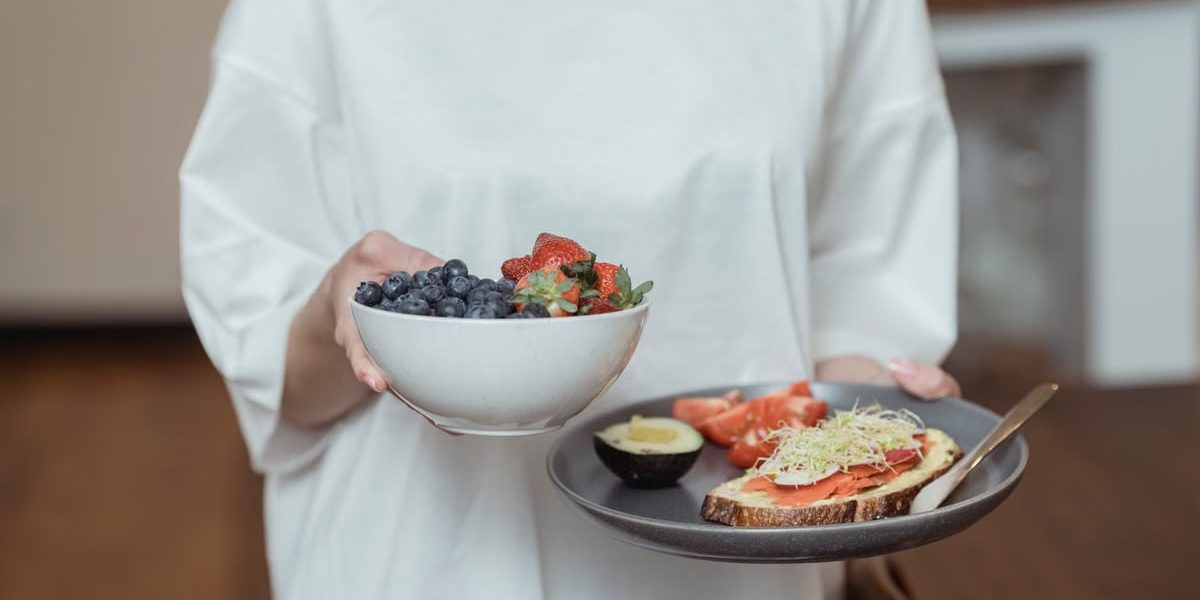 Person holding a bowl of fresh berries and a plate with avocado toast, displaying a healthy breakfast spread.