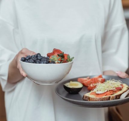 Person holding a bowl of fresh berries and a plate with avocado toast, displaying a healthy breakfast spread.