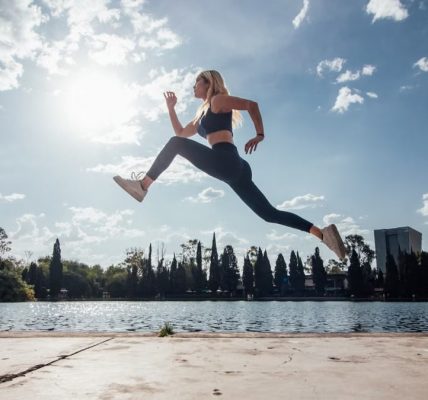 Woman performs an athletic jump by a lakeside in the park under a sunny blue sky.