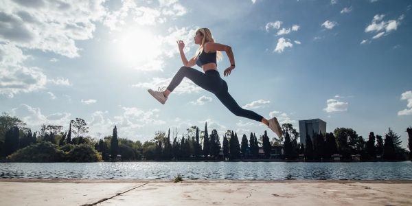 Woman performs an athletic jump by a lakeside in the park under a sunny blue sky.
